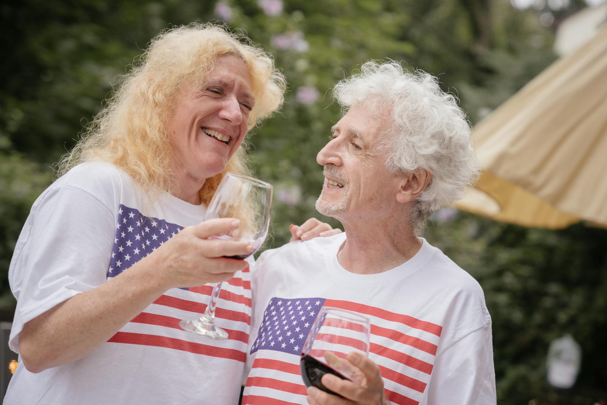 Elderly couple in American flag shirts enjoying wine together outdoors.