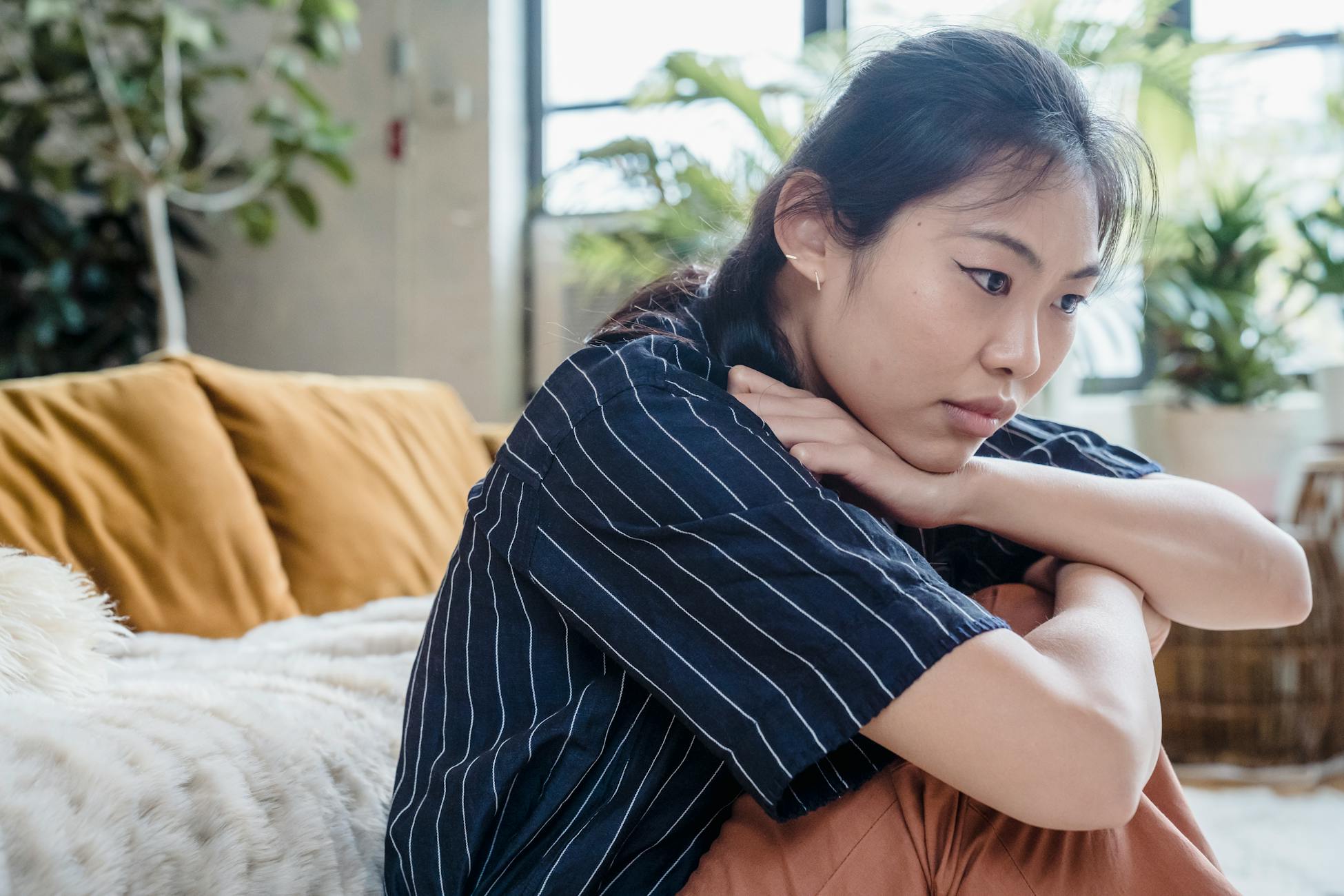 Young woman sitting pensively on a cozy couch indoors, surrounded by plants. Thoughtful and contemplative mood.