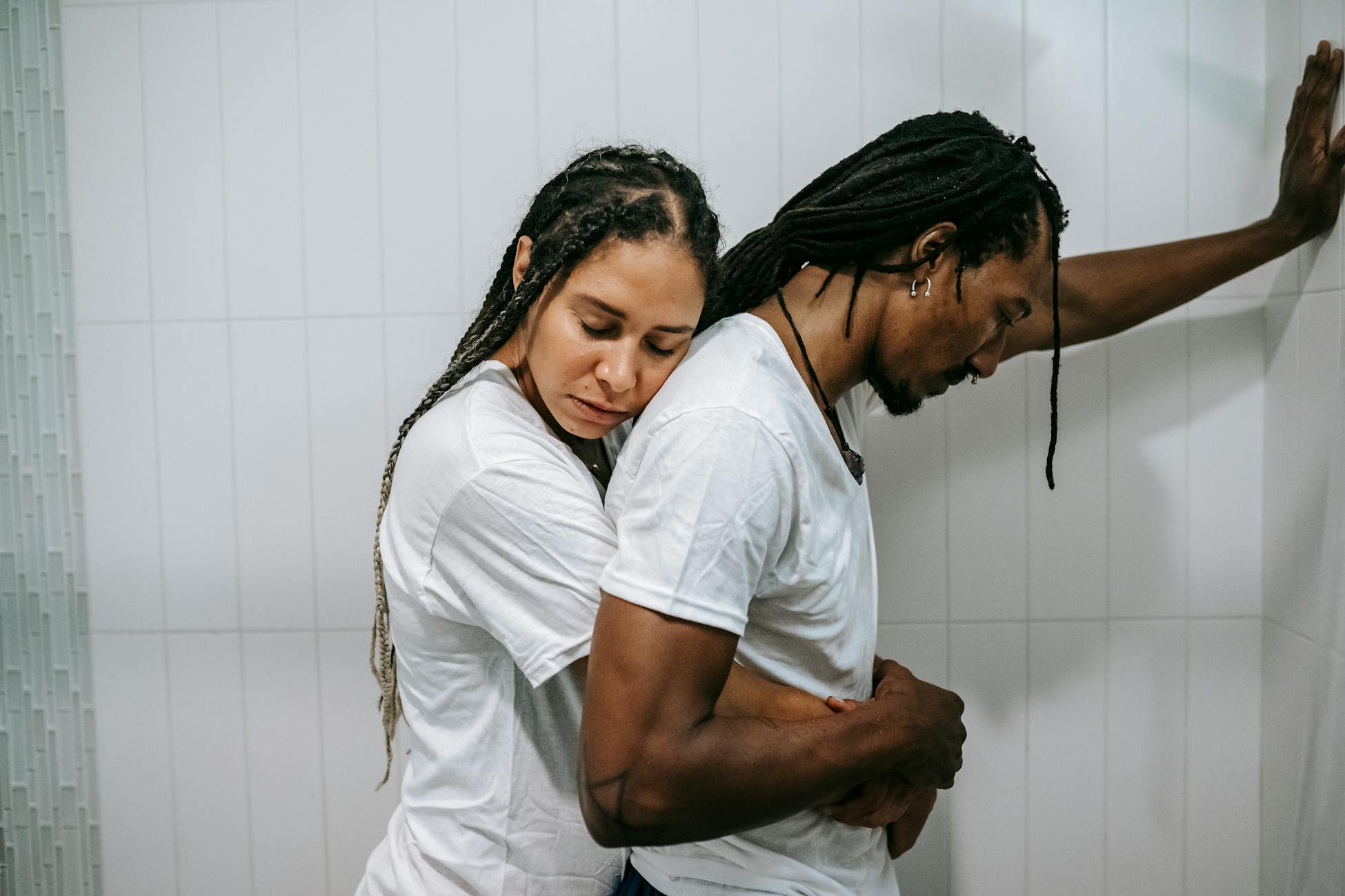 Side view sad loving African American female in white shirt standing behind and embracing depressed unhappy husband in bathroom