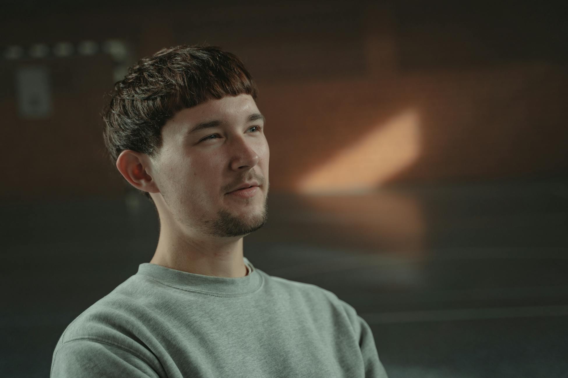 A man with a thoughtful expression sitting indoors, illuminated by soft natural light.