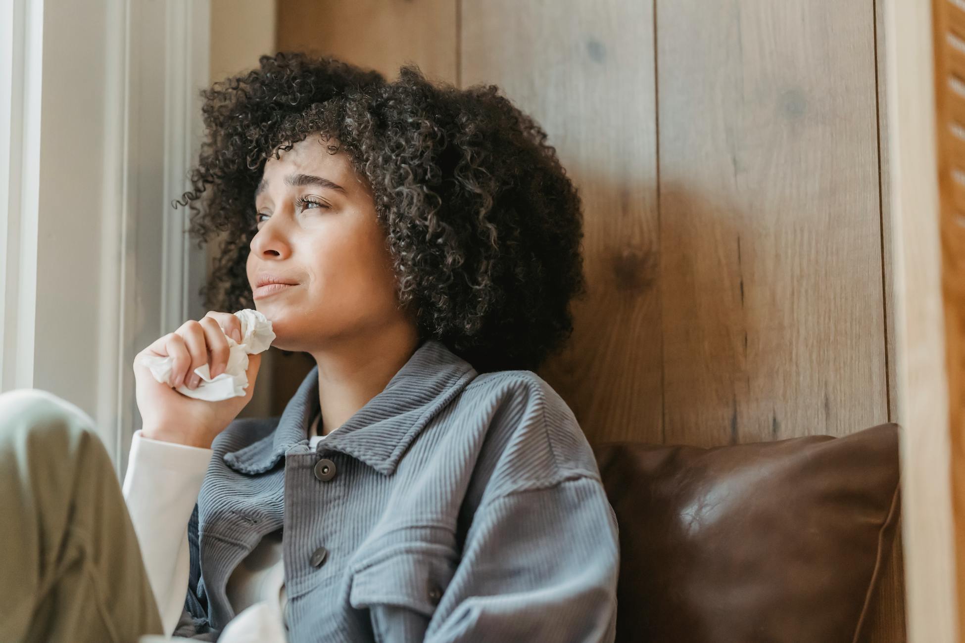 Young woman with curly hair in deep thought sitting by a window, conveying emotions of contemplation.