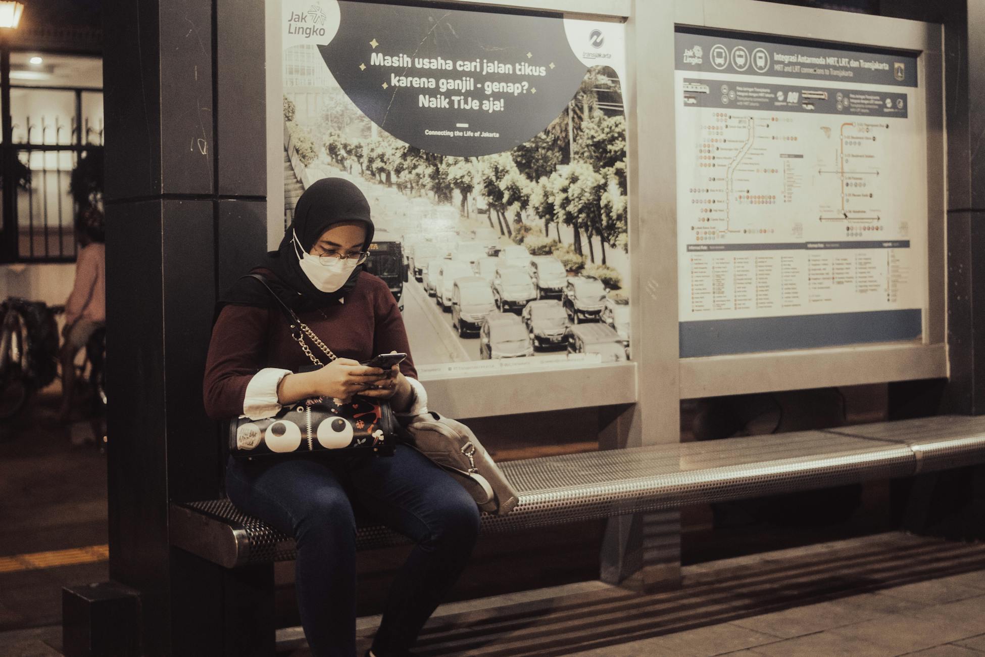 A woman wearing a mask and hijab uses her phone while sitting at a Jakarta bus stop at night.
