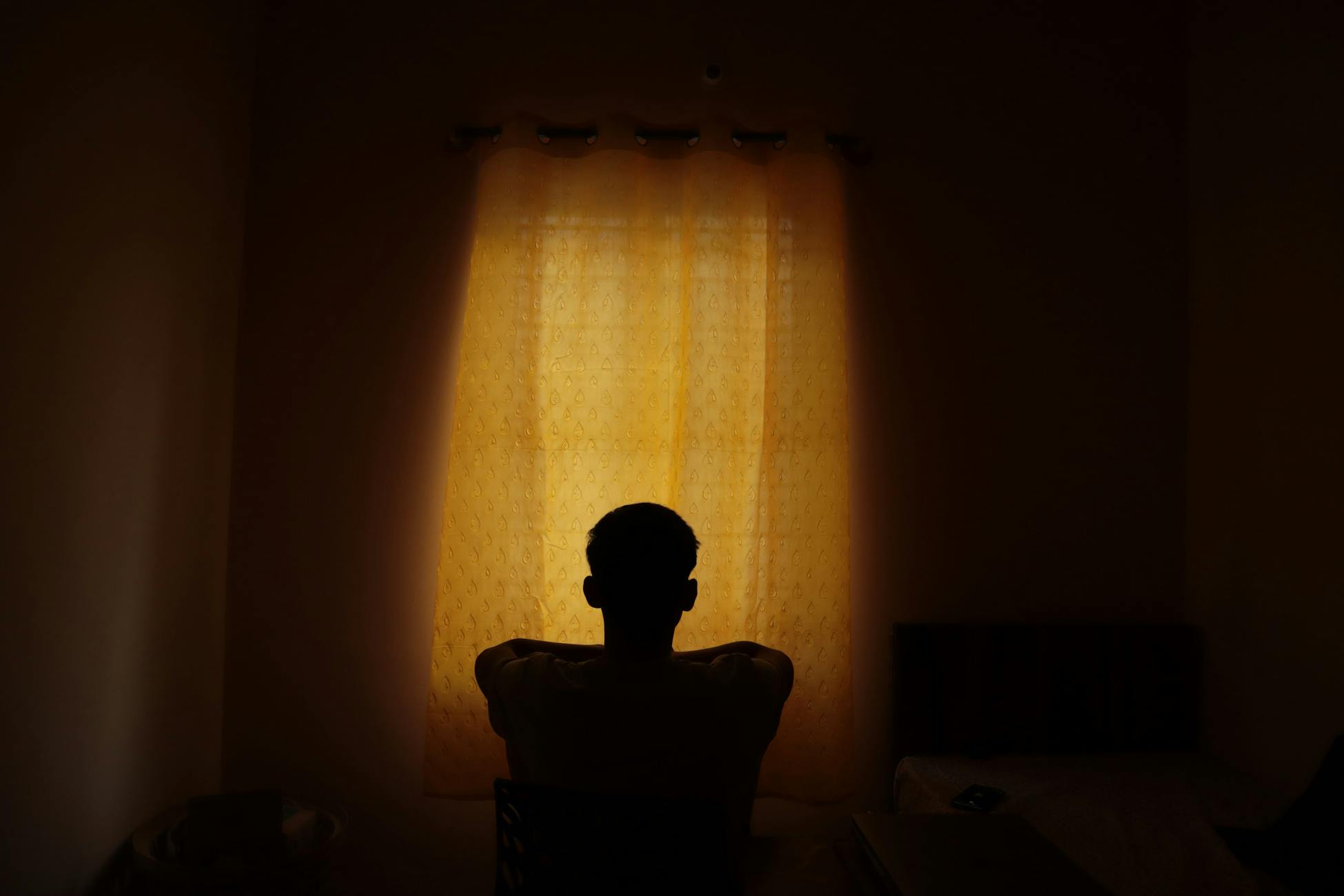 A backlit silhouette of a person sitting alone indoors, looking at a window with light filtered by curtains.