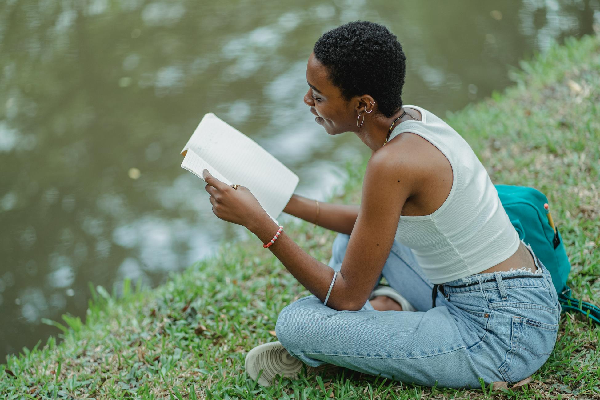 From above of positive young African American female student wearing trendy jeans sitting on grassy bank of lake and reading textbook