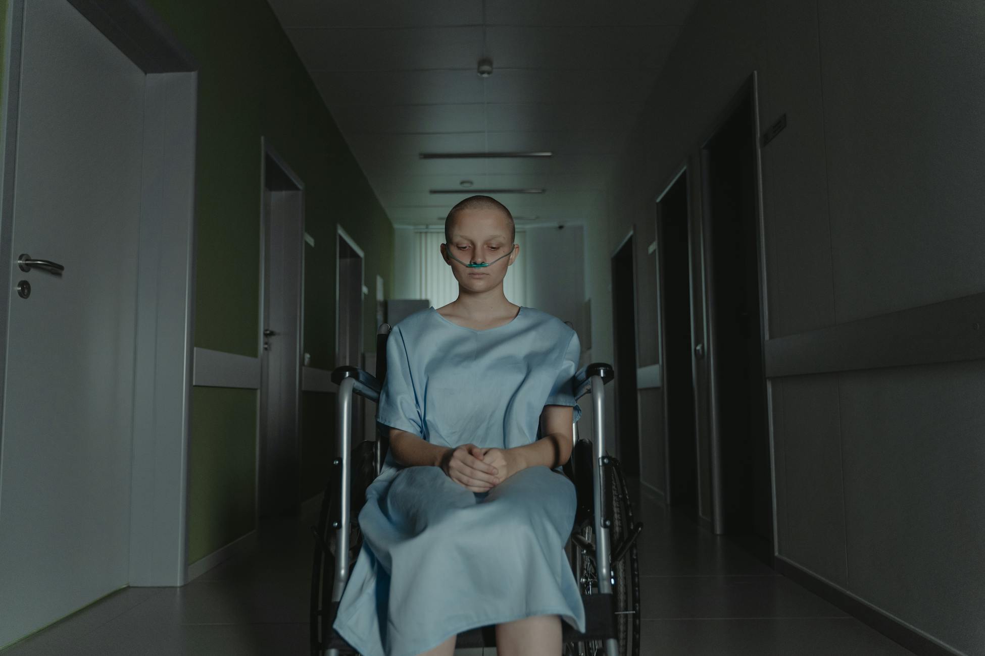 Woman in hospital gown sitting in wheelchair, deep in thought, in dimly lit corridor.