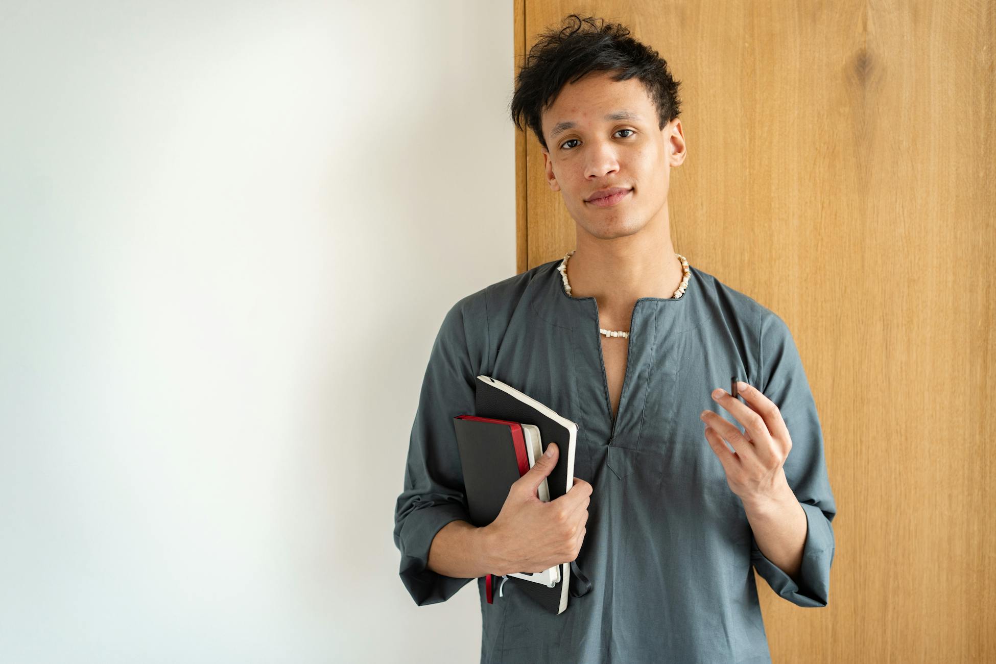 A young adult stands indoors holding notebooks against a wooden wall, exuding casual confidence.