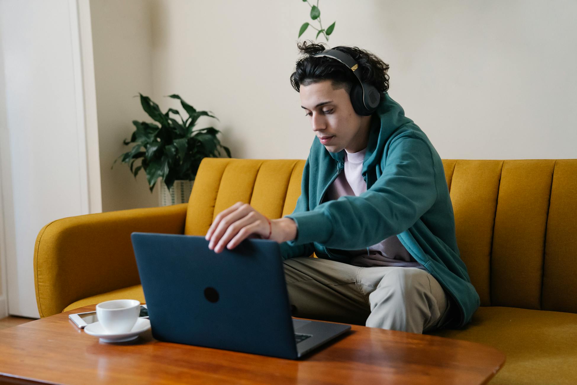 A young man wearing headphones using a laptop while sitting on a cozy sofa in a living room.