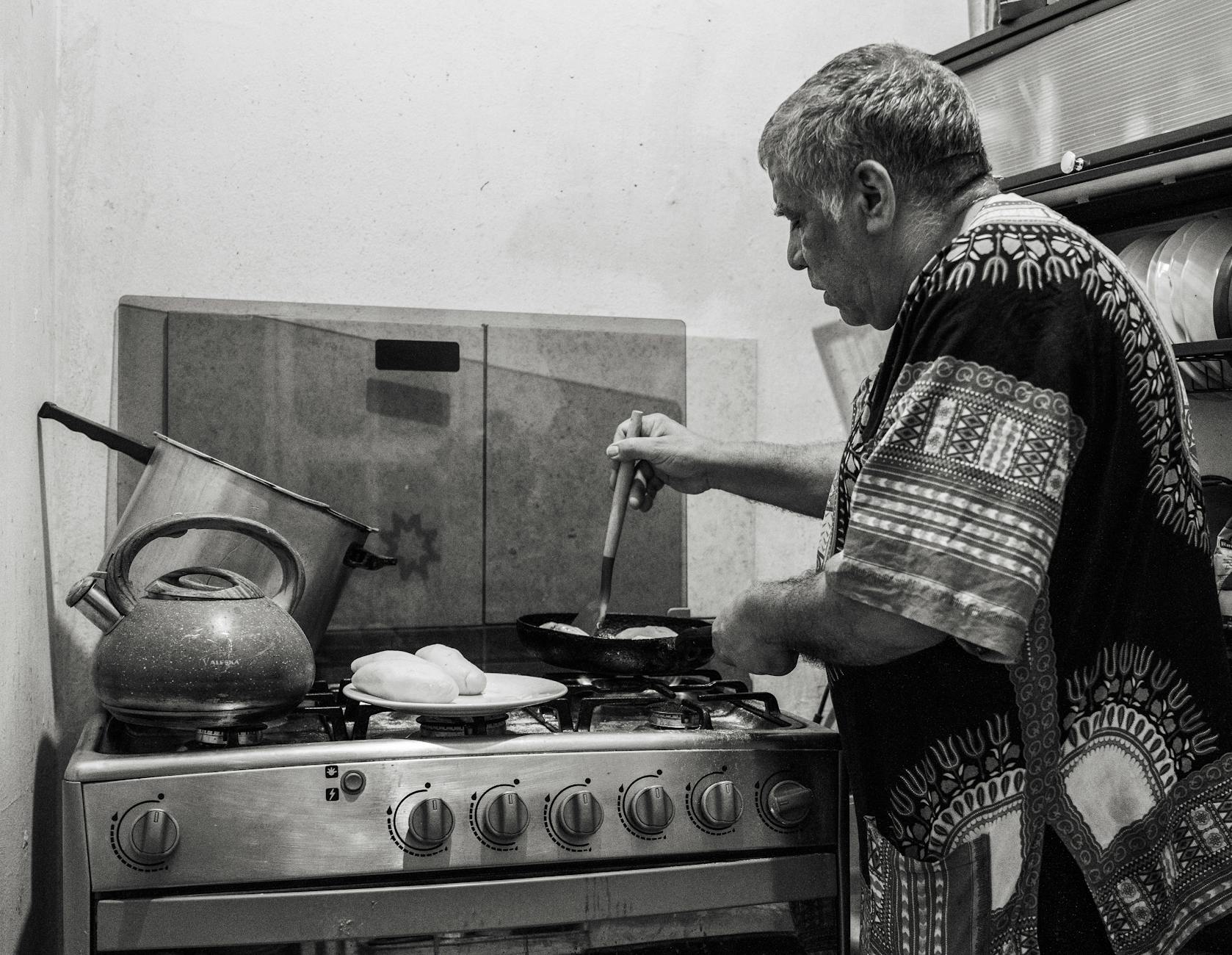 An elderly man cooking on a stove in a traditional kitchen setting, black and white photo.