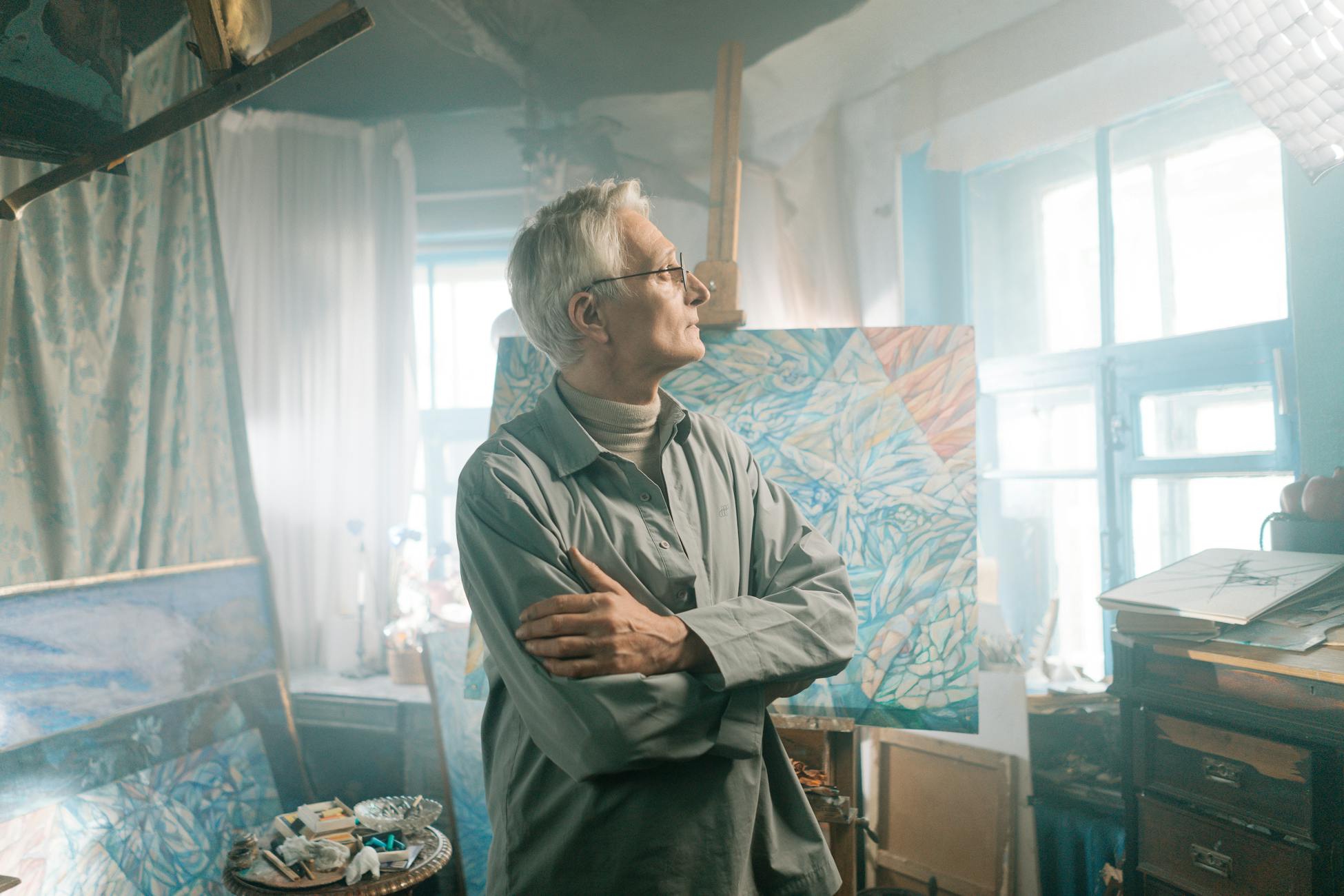 Senior man stands in an artistic studio, surrounded by paintings and natural light, contemplating his work.