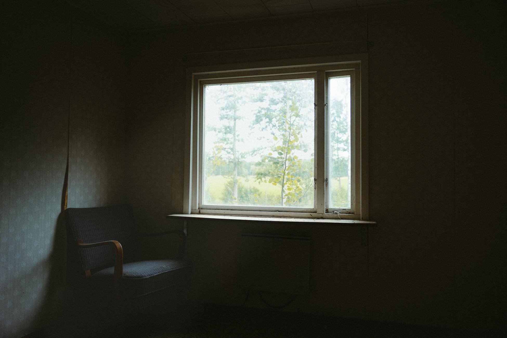 Dark room interior with a chair and window overlooking a tranquil outdoor scene.