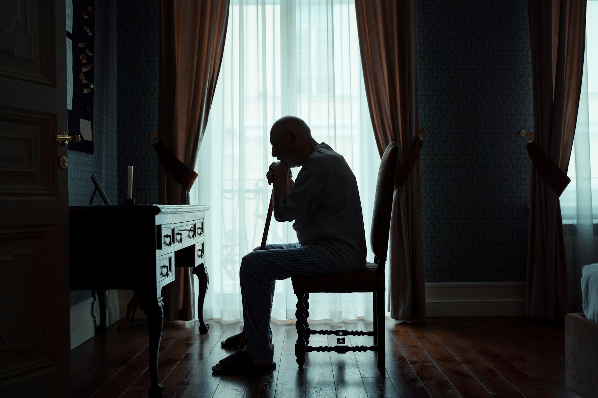 Silhouette of an elderly man sitting indoors by a window, contemplating life.