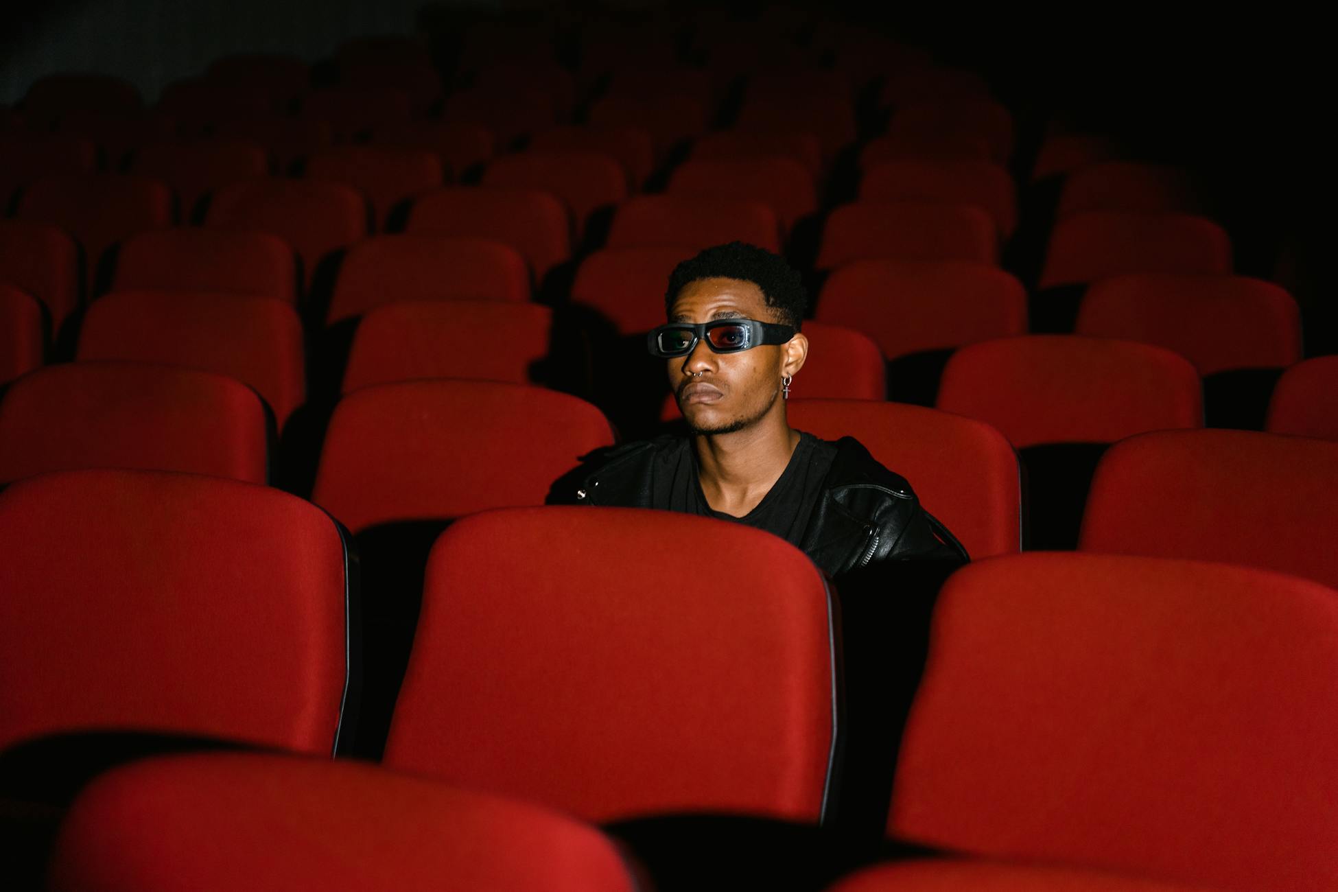 A man wearing 3D glasses sits alone in a movie theater with red seats, creating an isolated atmosphere.