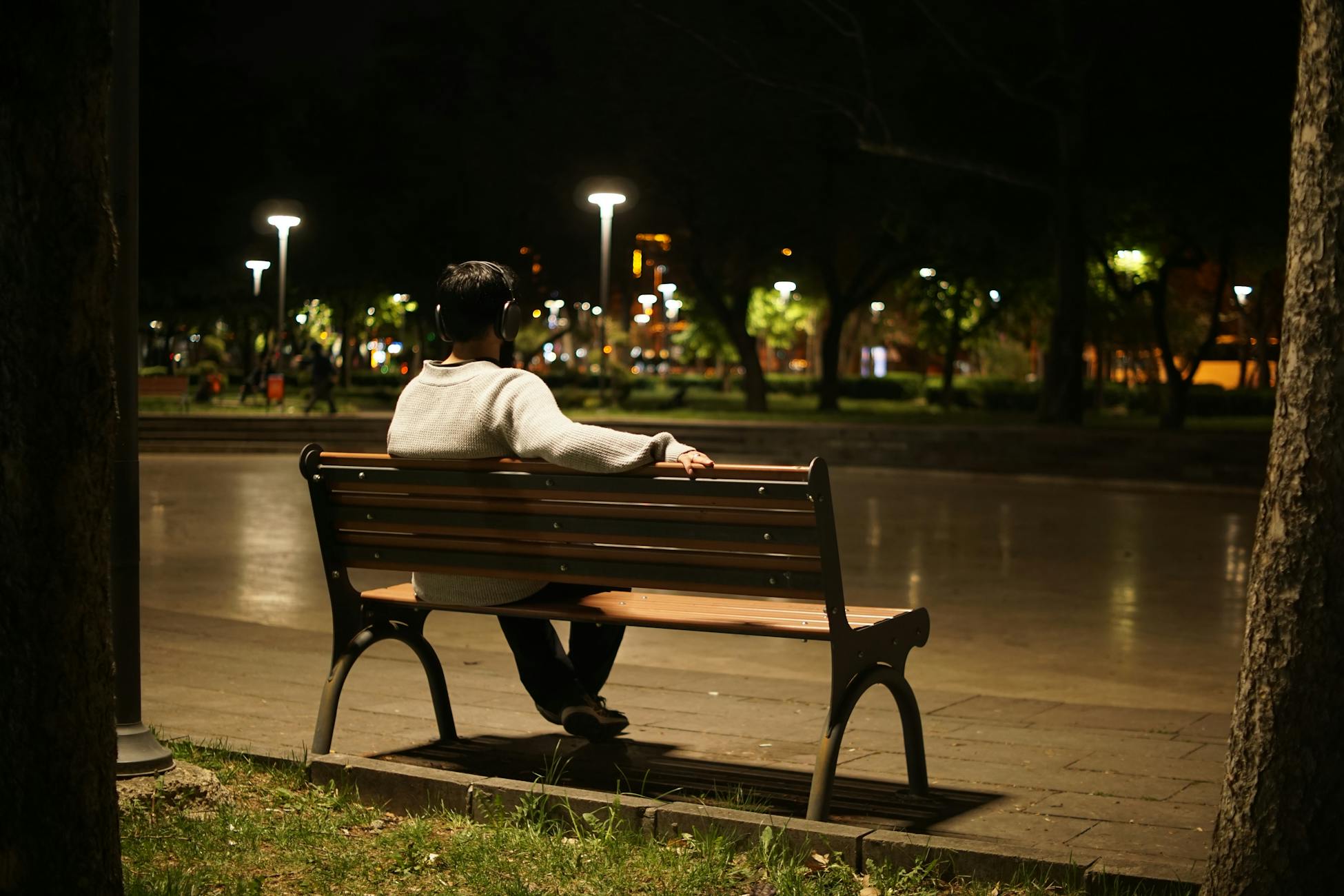 Solitary figure listening to music on a park bench under night streetlights, urban solitude.