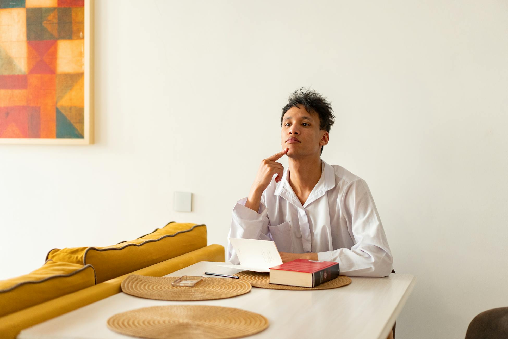 Man in white shirt pondering at a dining table with books, showcasing modern interior.