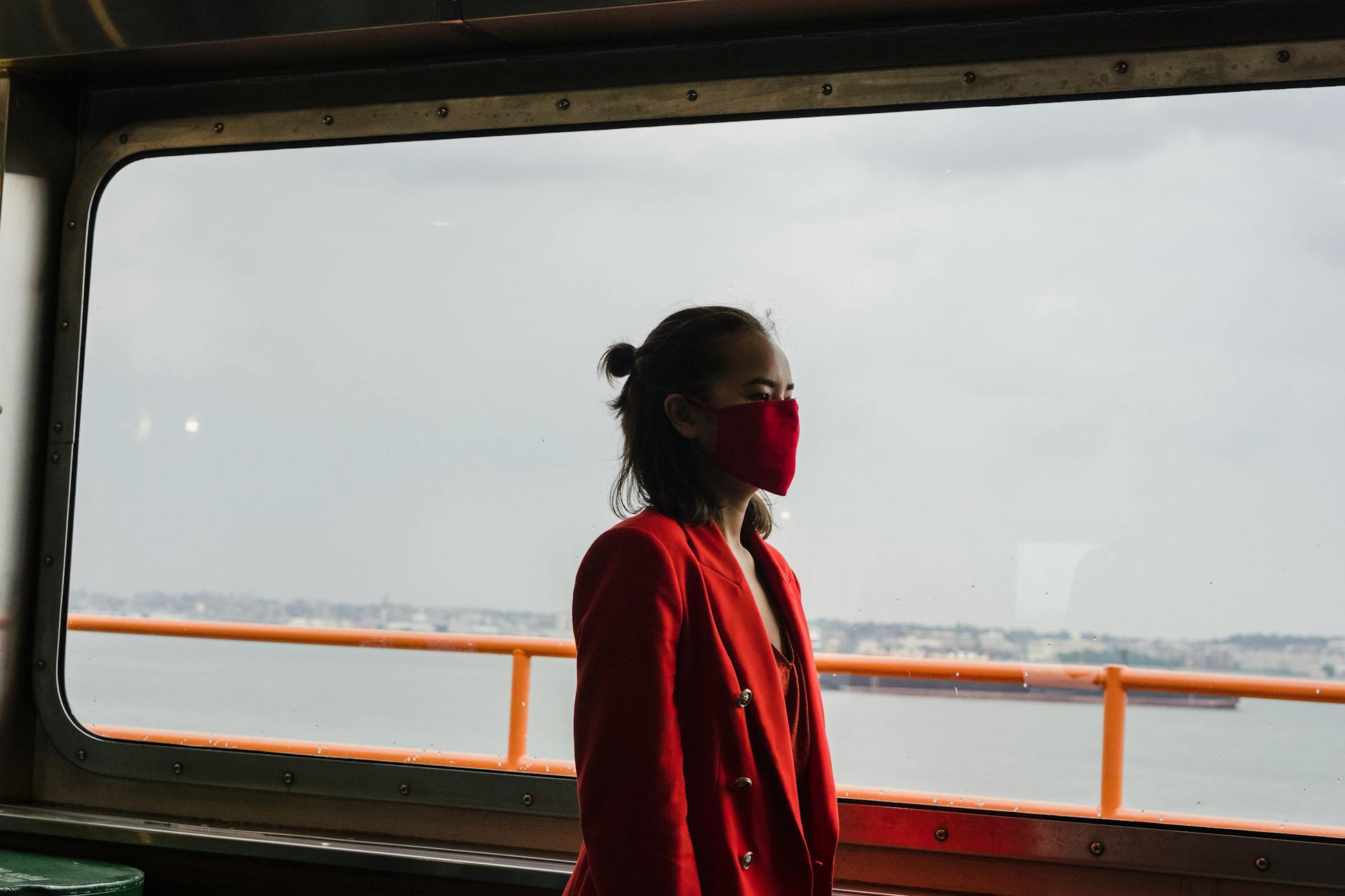 A woman in a red suit and face mask stands by a window overlooking a waterfront.