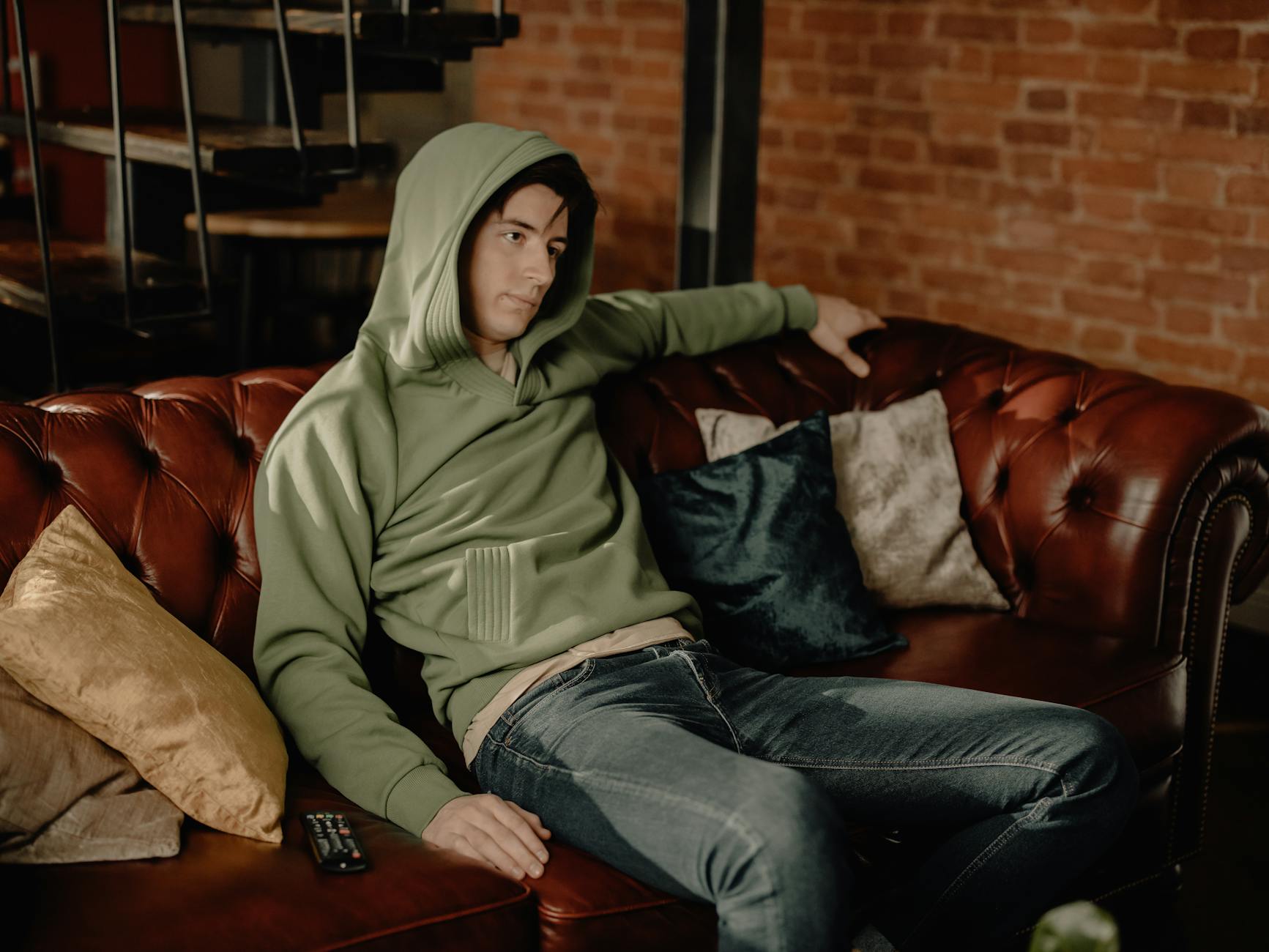 Young man in hoodie relaxing on leather sofa in cozy home interior, feeling contemplative.
