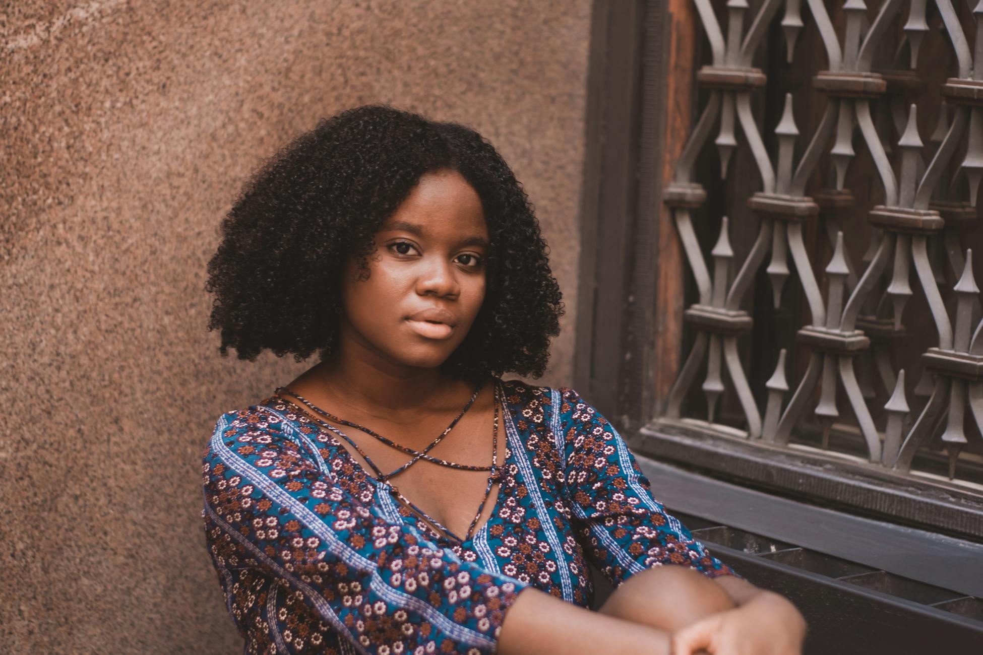A woman with curly hair in casual wear sitting against a decorative window, captured during a photoshoot.