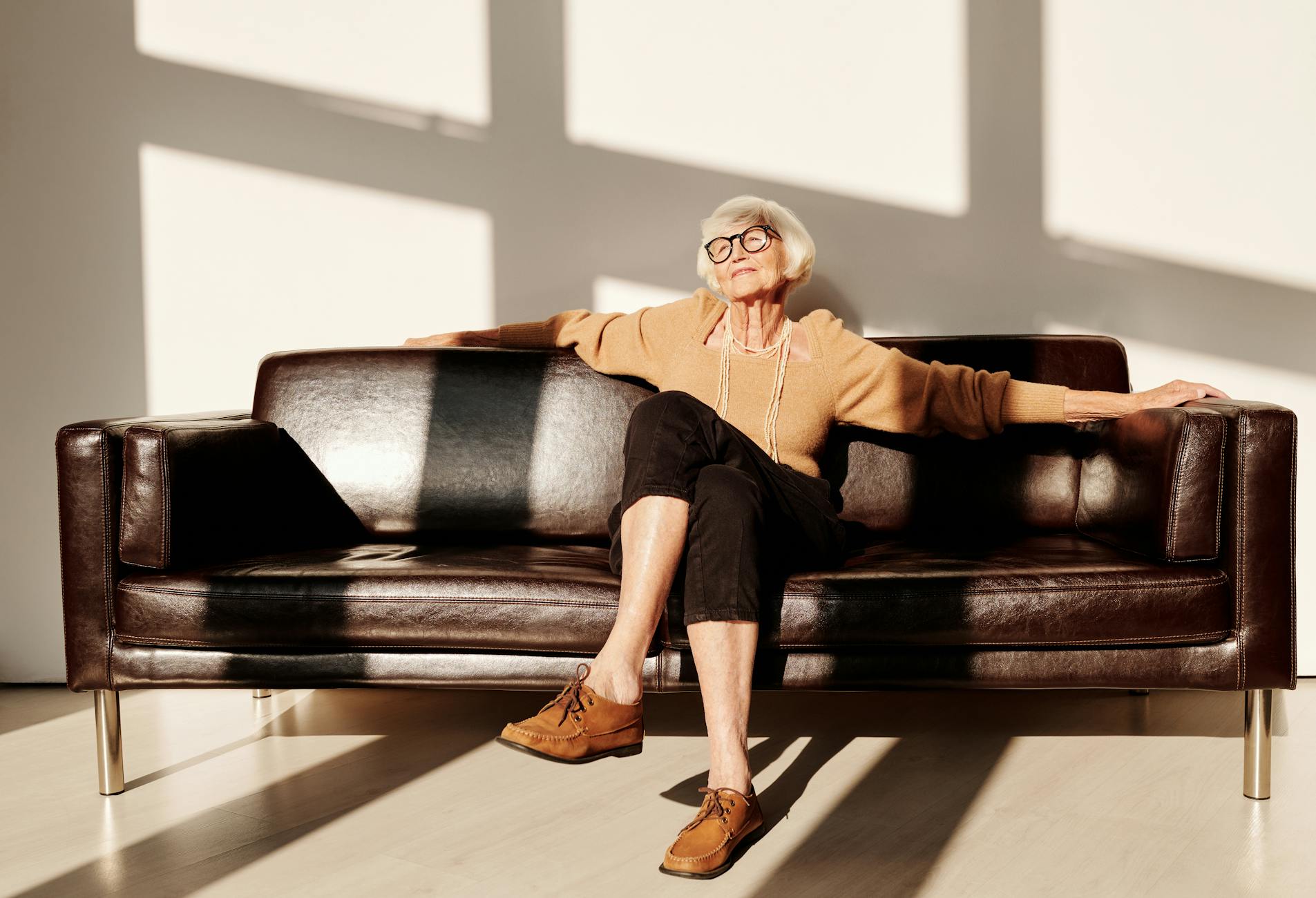 Elderly woman with gray hair and eyeglasses relaxing on a leather sofa in a bright room.