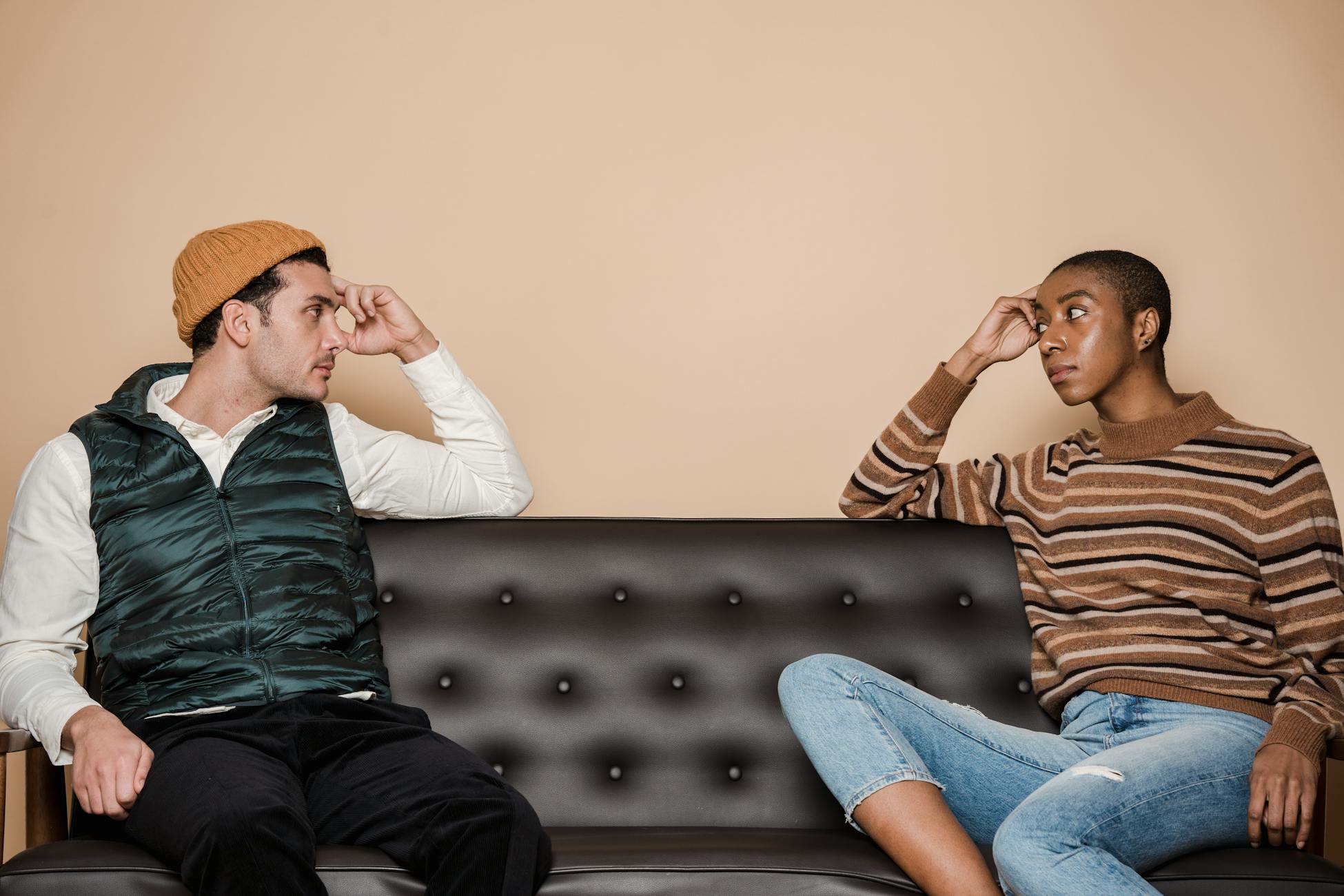 Diverse couple in casual clothes looking at each other with serious faces while sitting on sofa during quarrel in living room