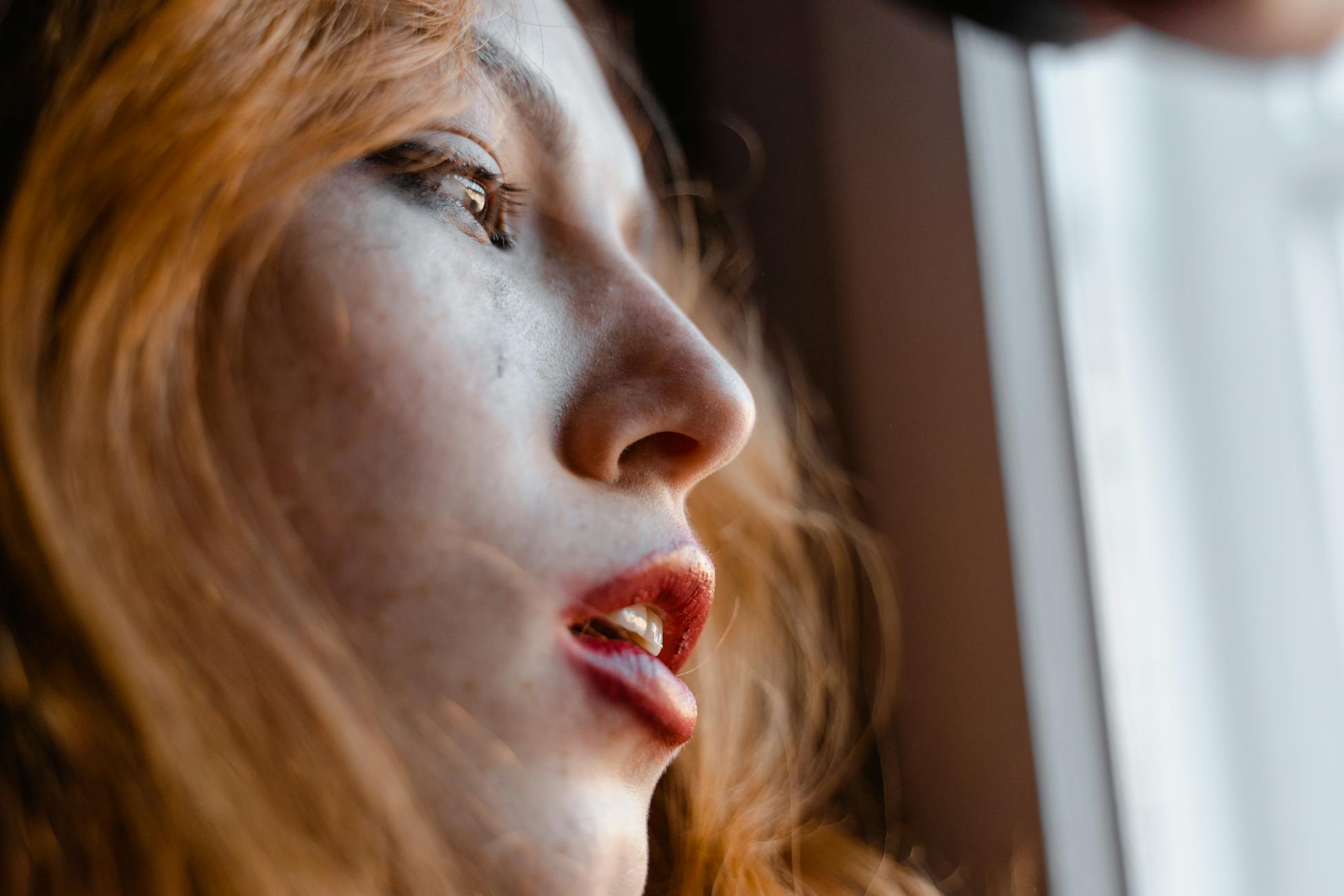 A young woman with red hair looks thoughtfully out a window, capturing an emotional moment.