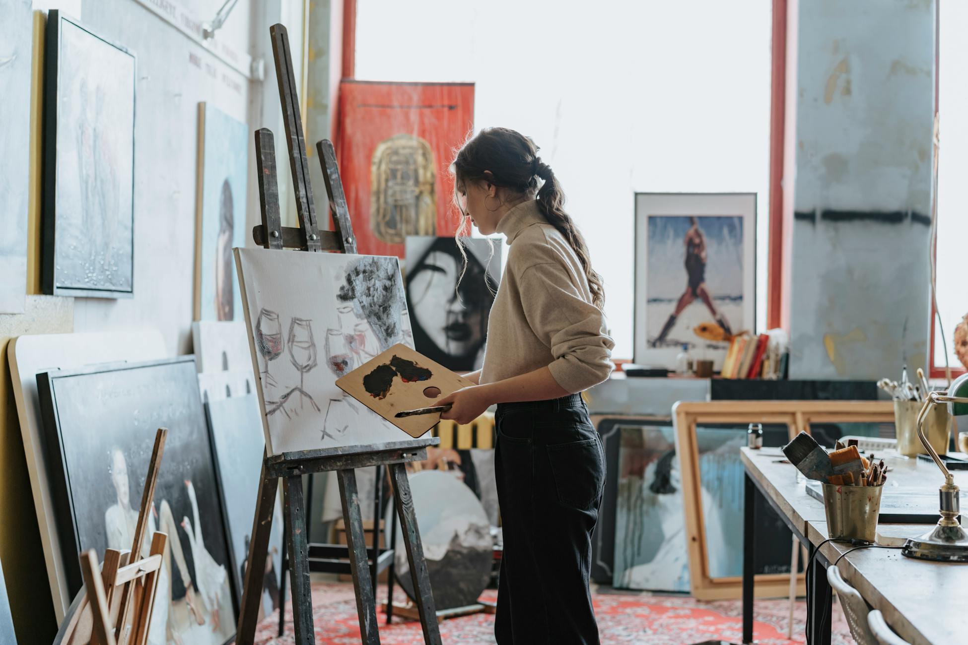 A woman artist works on a painting in a creative studio space filled with art and light.