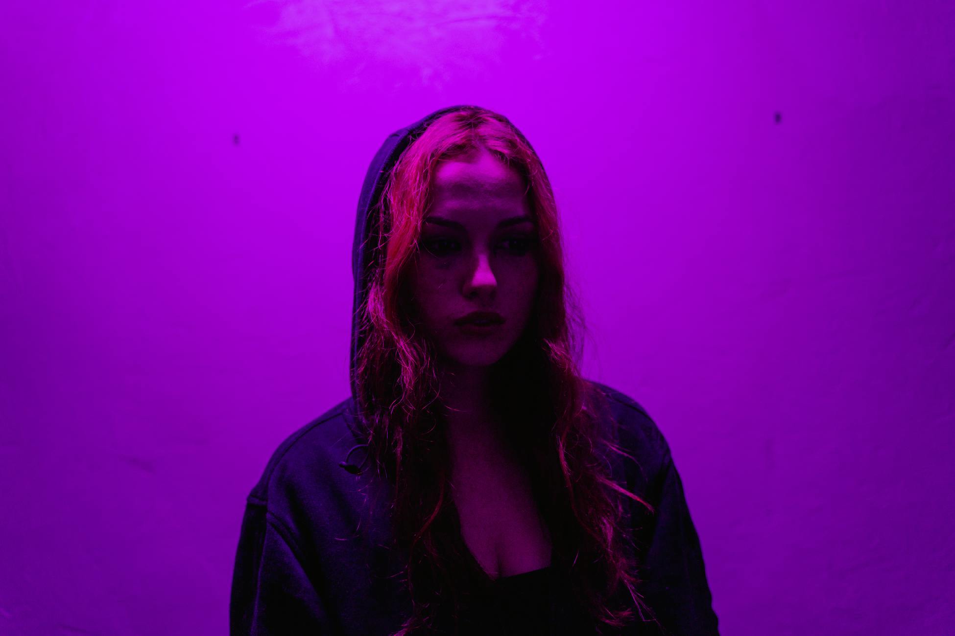 A woman in a hoodie stands under dramatic purple lighting, evoking emotion.