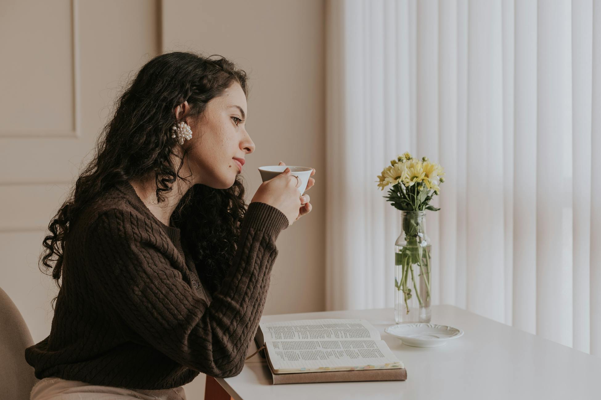 A woman relaxes with tea and a book by a bright window, embracing a peaceful moment.
