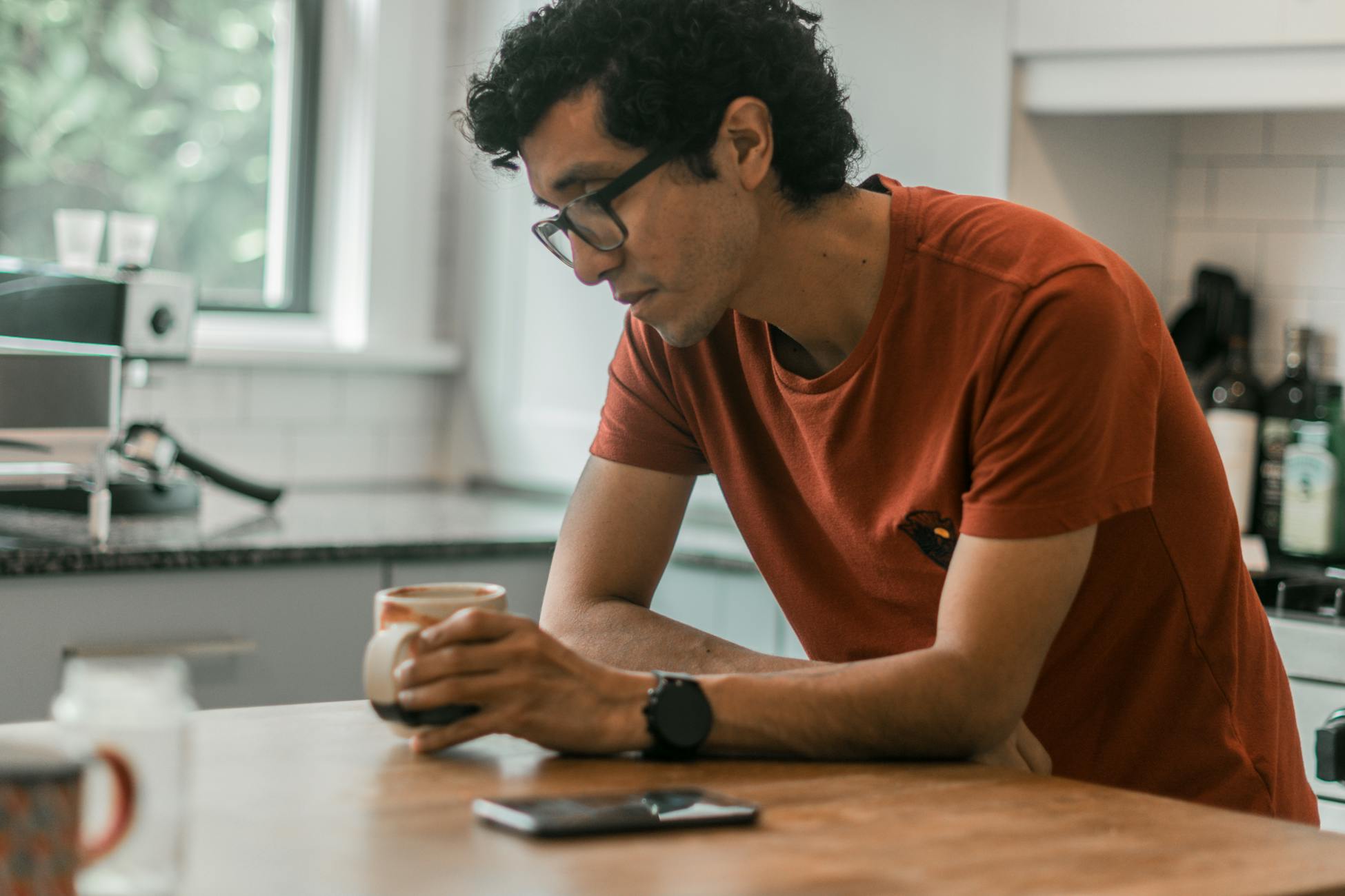A man in a red t-shirt leans on a kitchen counter, holding a coffee mug and looking thoughtful.