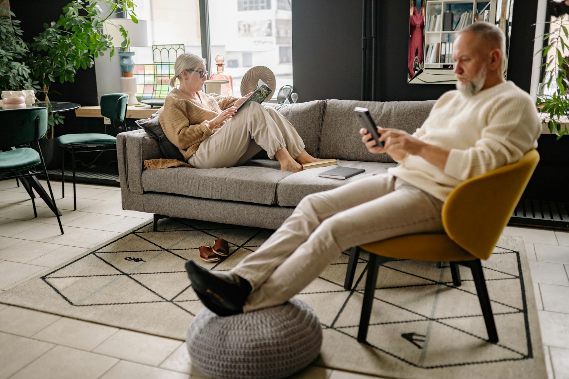Senior couple relaxing indoors, reading and using a smartphone in a cozy living room.