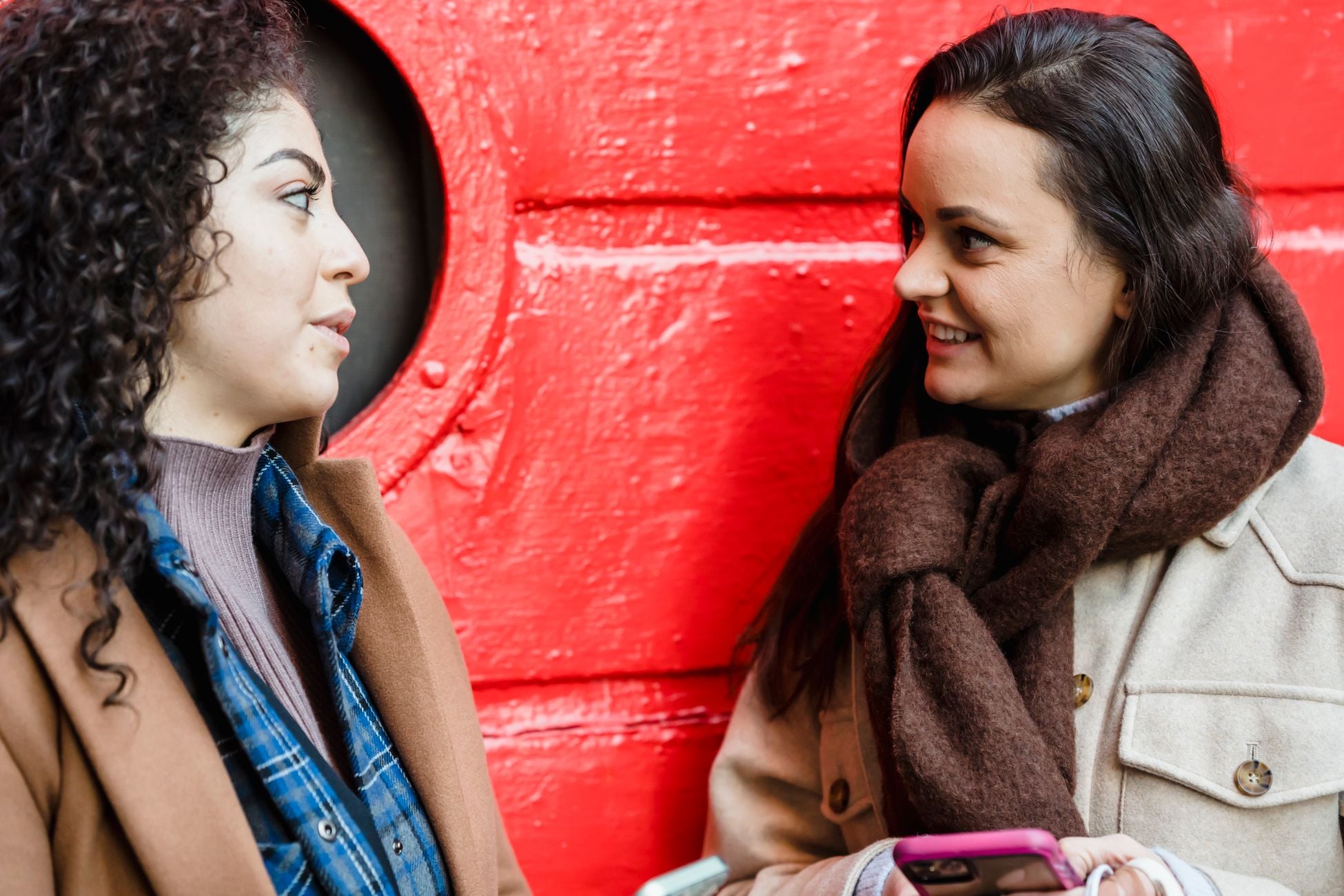 Positive young multiethnic female friends in casual outfits standing on street near red metal door and chatting